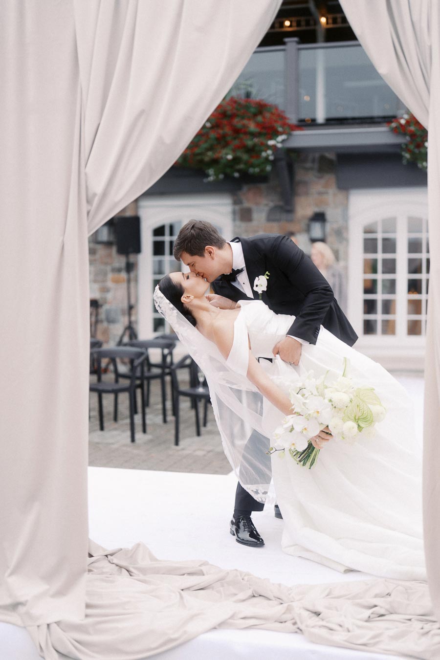 Bride and groom sharing a romantic kiss under draped curtains at an outdoor wedding ceremony, with the bride holding a bouquet of white flowers.