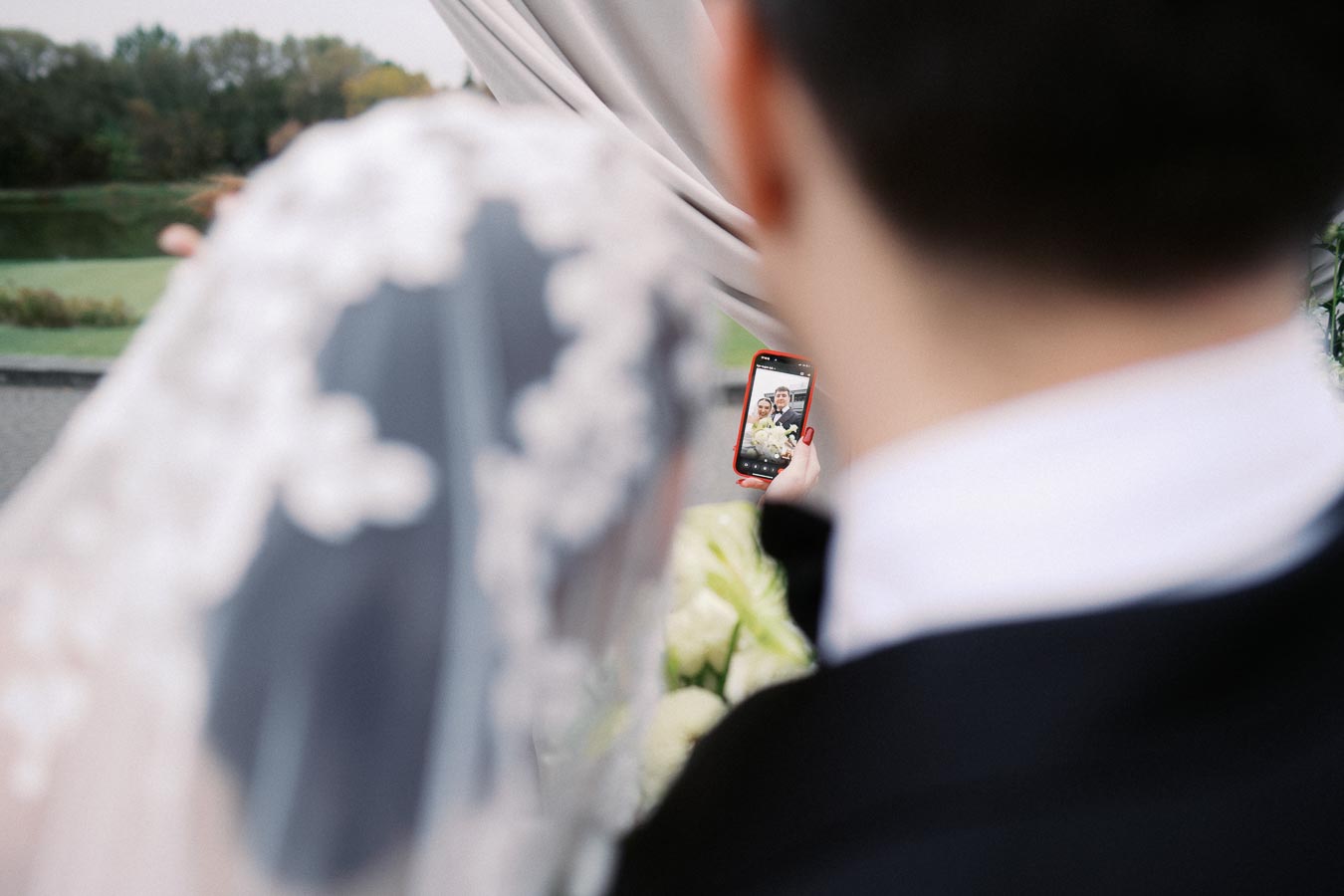 Bride and groom taking a selfie at an outdoor wedding ceremony, showcasing a close-up of their smartphone capturing the moment, with a bridal veil and floral bouquet visible.
