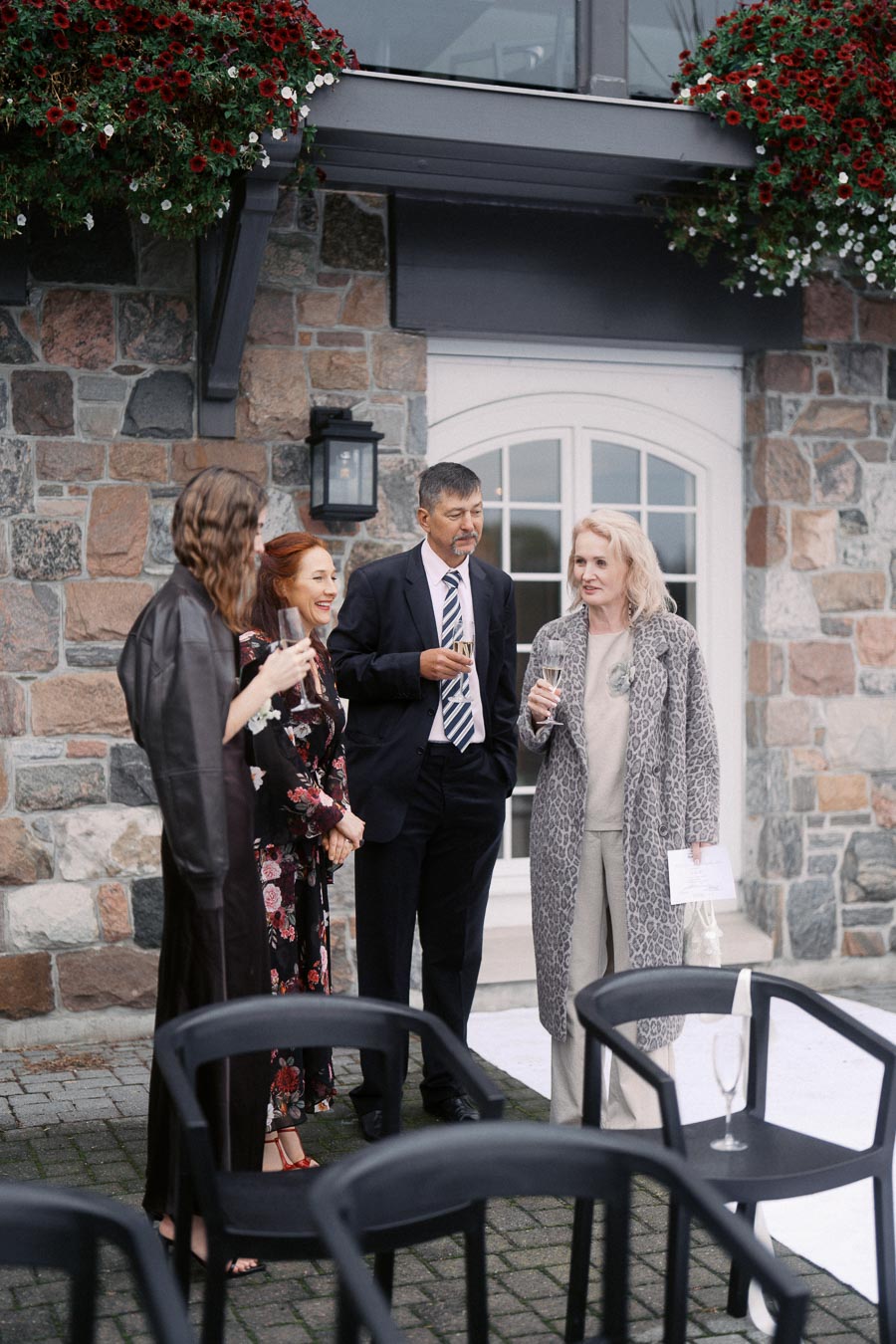 Group of elegantly dressed people socializing outdoors at a formal event, standing in front of a stone building with flowering plants.