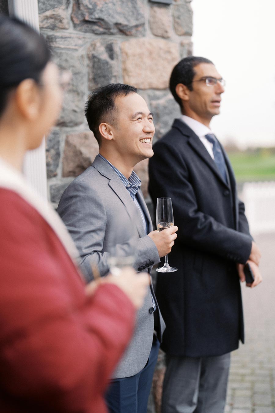 Three people in business attire enjoying a social gathering, holding champagne flutes, and standing outside near a stone wall.