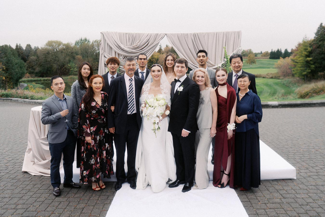 A joyful group photo of a bride and groom surrounded by friends and family at an outdoor wedding ceremony. The couple stands in front of an elegant draped archway, set against a scenic backdrop of greenery and trees, creating a picturesque wedding scene. Everyone is dressed in formal attire, with smiles and celebratory expressions.