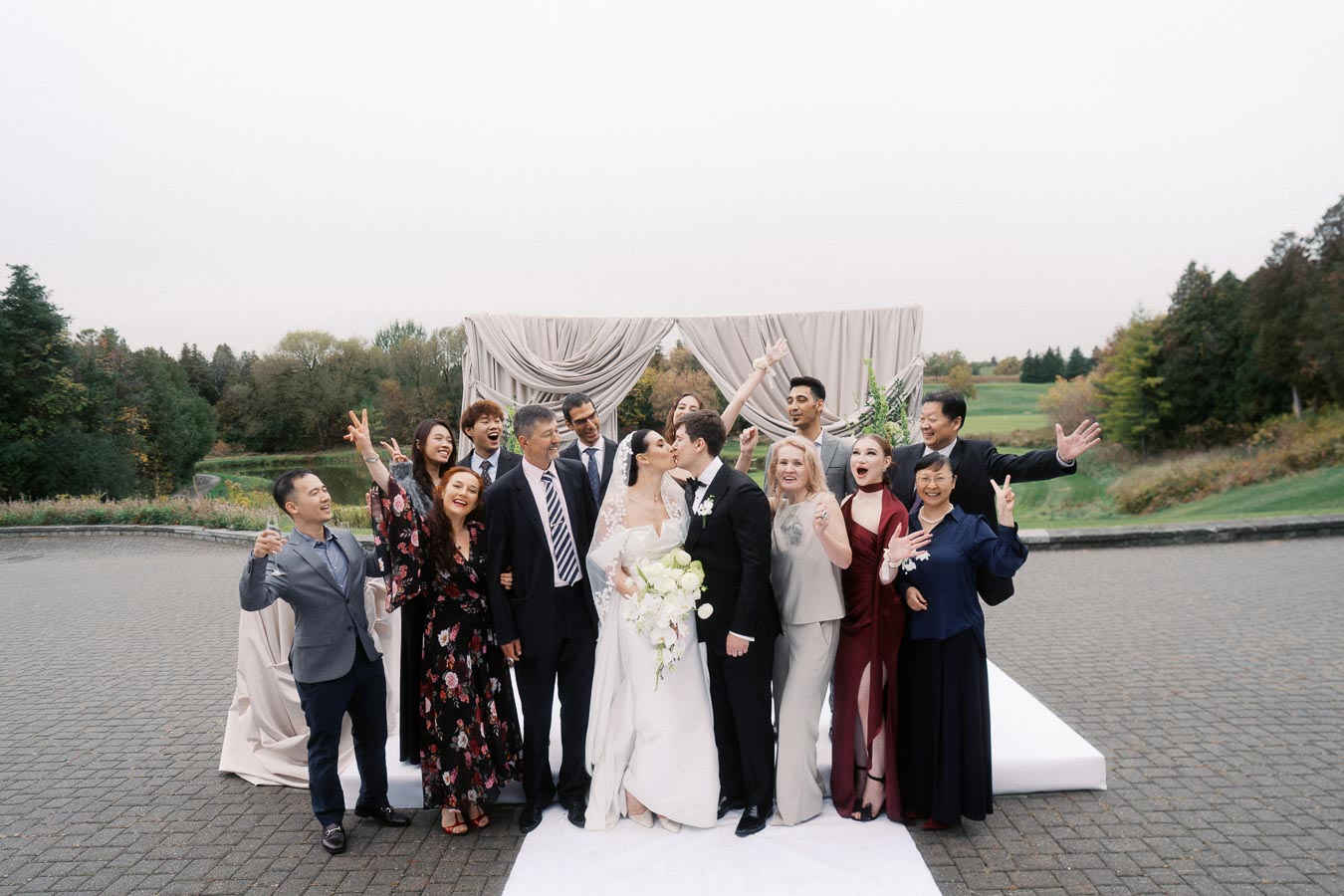 A joyful wedding group photo with a bride and groom sharing a kiss in the center, surrounded by smiling guests in formal attire, captured outdoors against a scenic backdrop with greenery and trees.