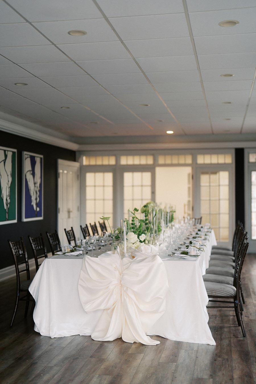 Elegant event hall with a long table set for a formal dinner, adorned with white tablecloths, floral centerpieces, and decorative bows.