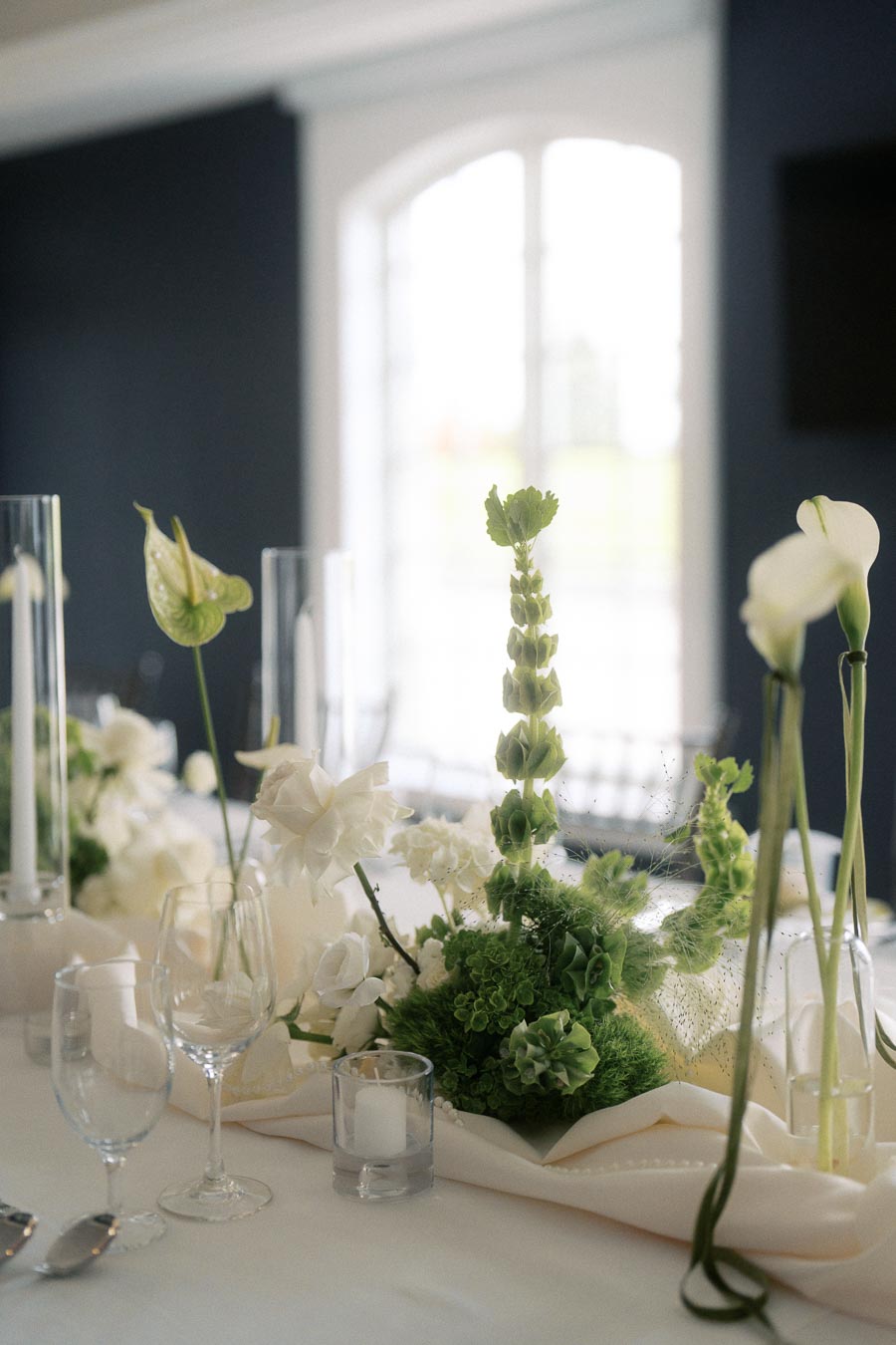 Elegant wedding table setting with a sophisticated floral centerpiece featuring green and white flowers, surrounded by glassware, candles, and soft natural light from a large window in the background.