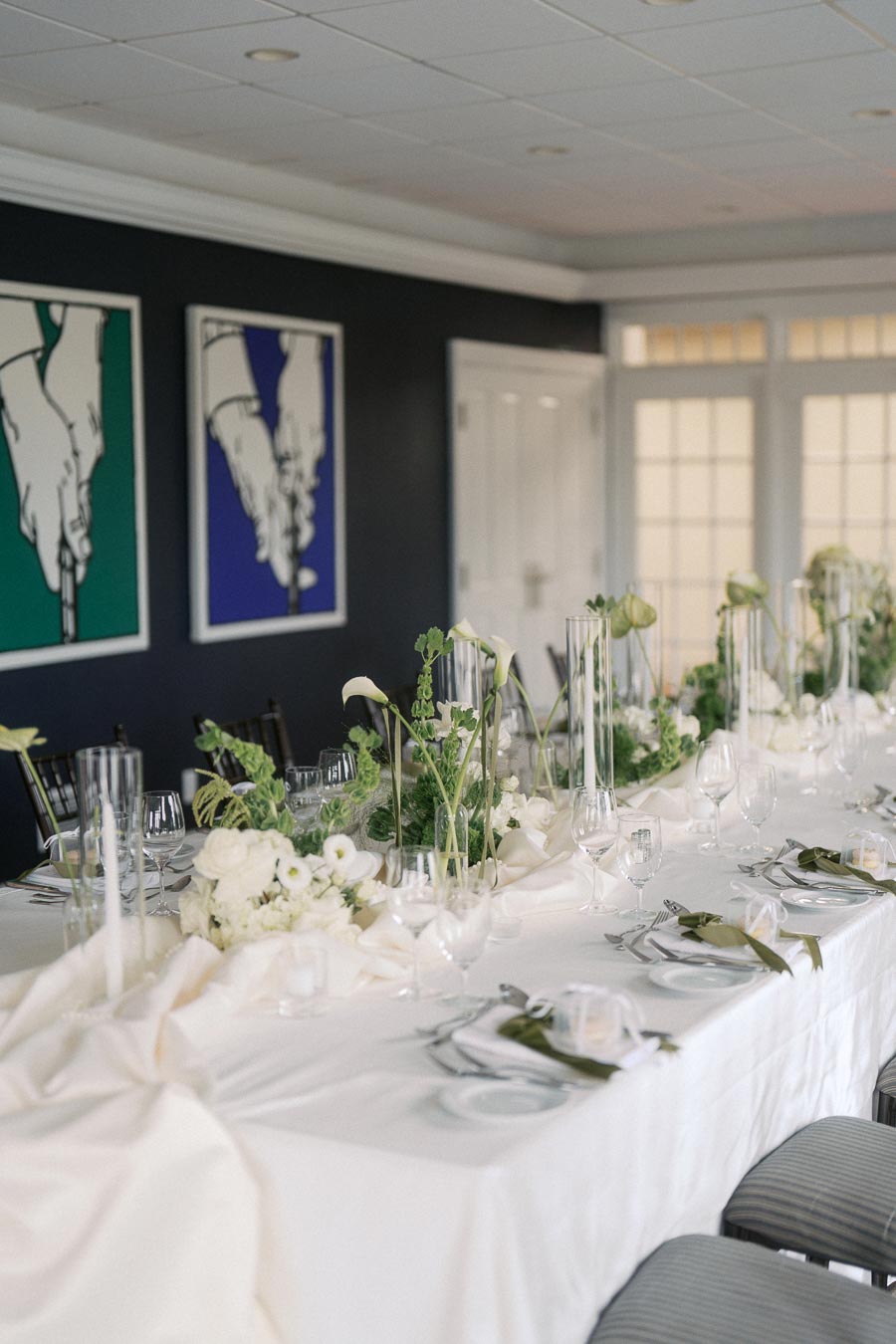 Elegant dining table setup with white linens and floral centerpieces featuring green and white flowers, in a sophisticated room with modern art on the walls.