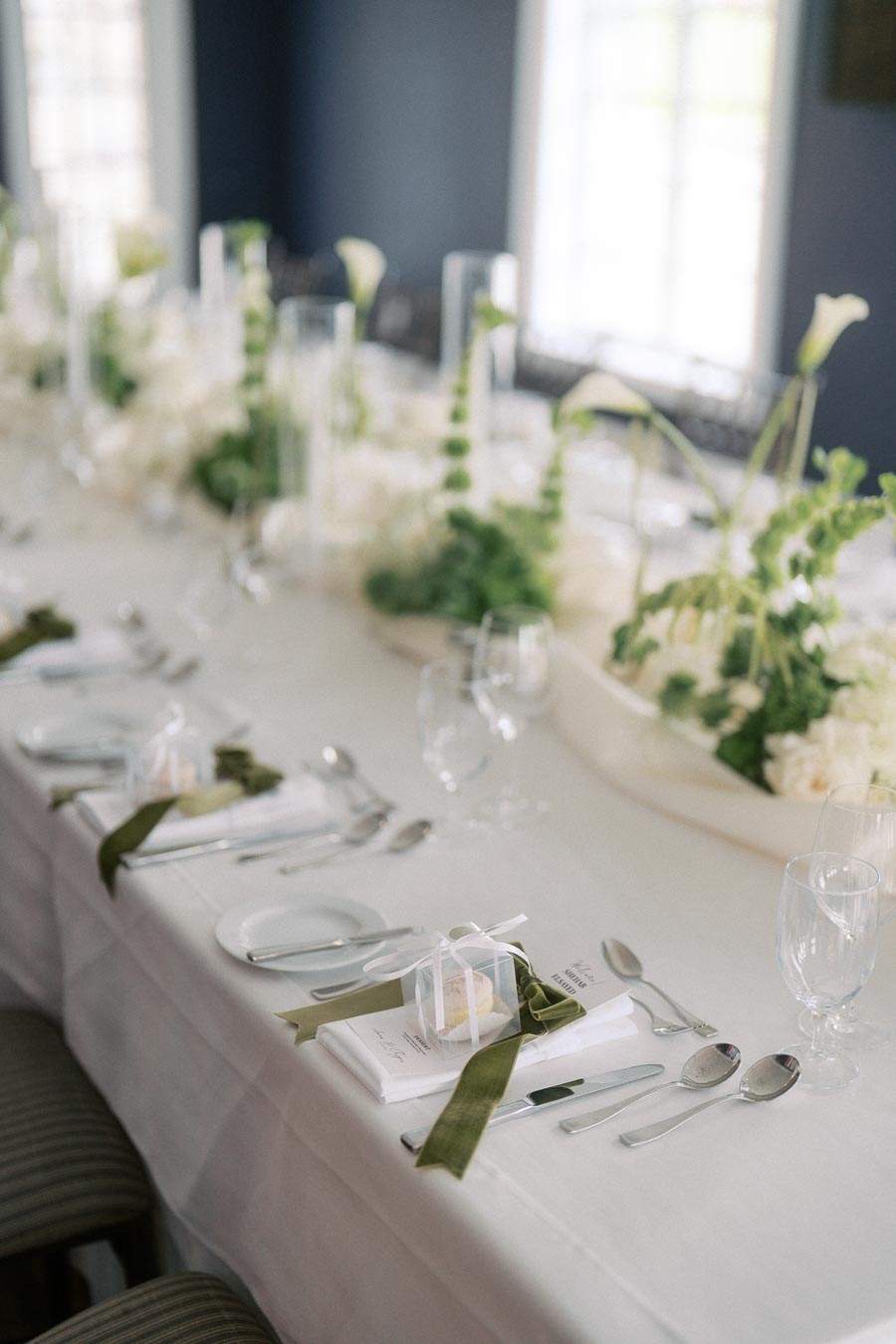 Elegant wedding table setting with white and green floral centerpieces, neatly arranged cutlery, and delicate gift boxes tied with green ribbons, set in a softly lit room.