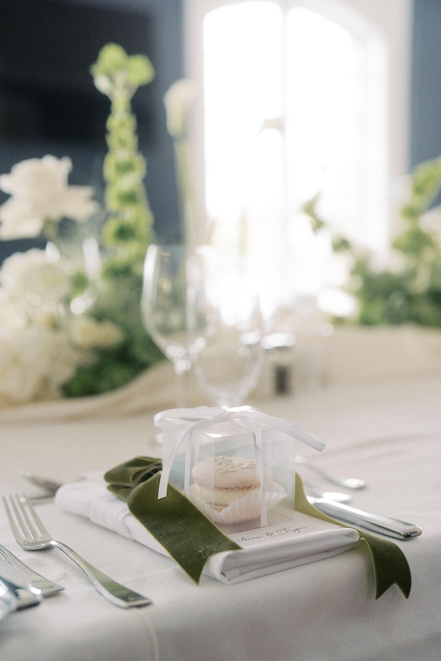 Elegant wedding table setting with white and green floral centerpiece, decorated with a sheer gift box containing a macaron, placed on a folded napkin tied with a green ribbon.