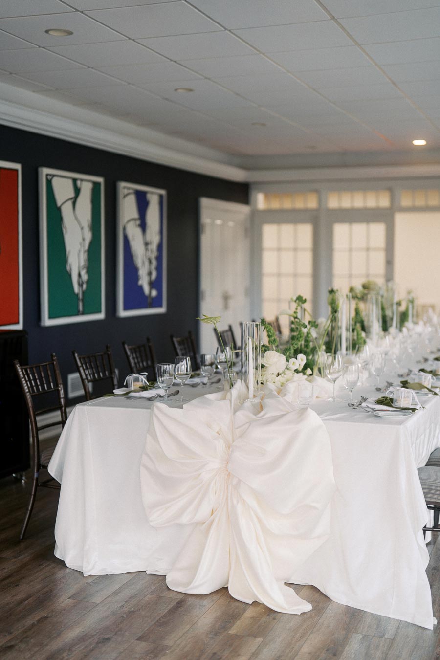 Elegant wedding reception table setting with white linen tablecloth, large decorative bow, and floral centerpieces, set within a modern room featuring colorful abstract artwork on the walls.