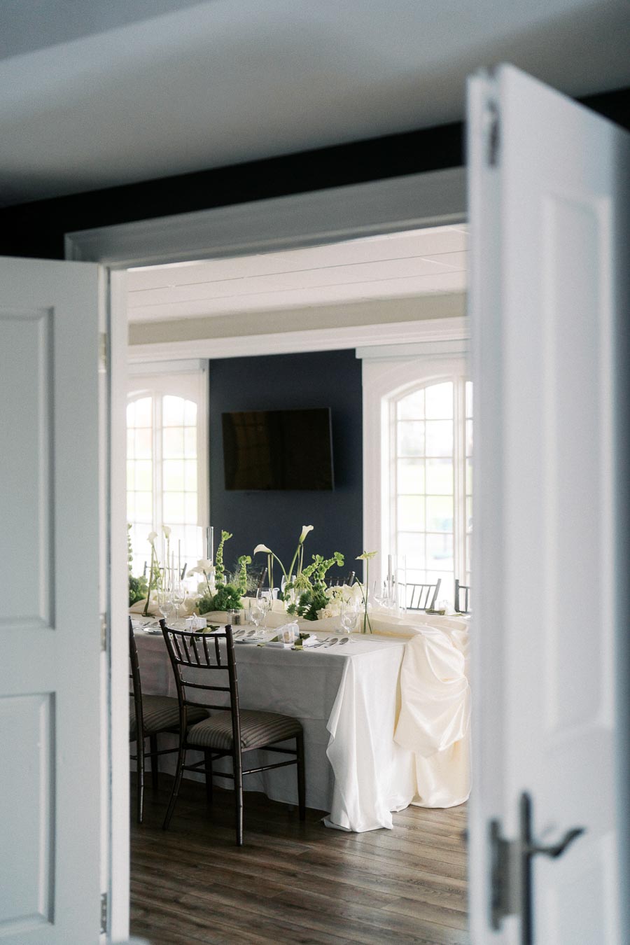 Elegant dining room setup with a white tablecloth, green floral centerpieces, and neatly arranged chairs captured through open double doors, ideal for a wedding or event.