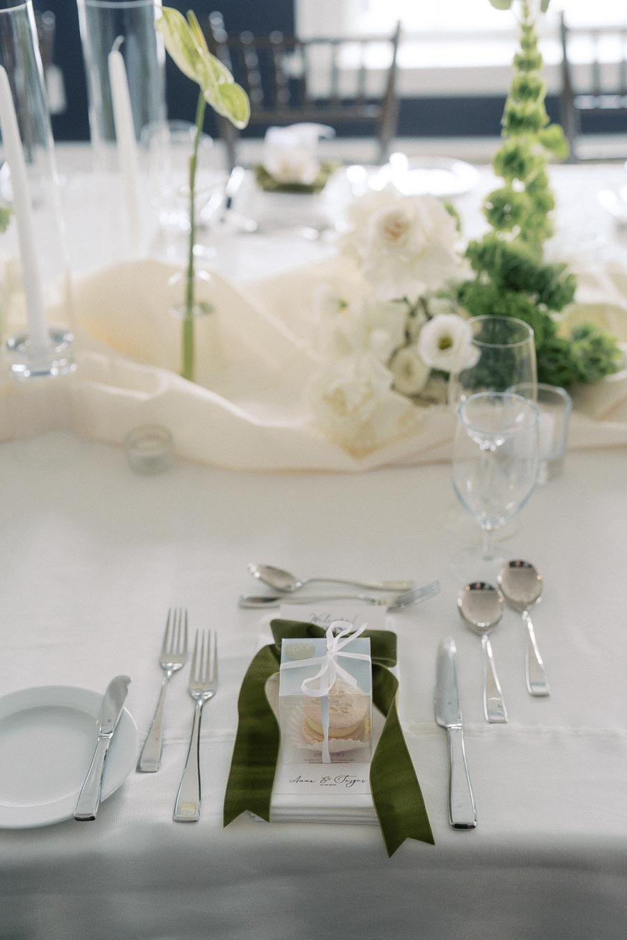Elegant wedding dining table with white tablecloth, featuring a neatly arranged setting of silver cutlery, clear wine glasses, and a small white gift box tied with a ribbon. Greenery and white flowers add a fresh touch to the centerpiece.