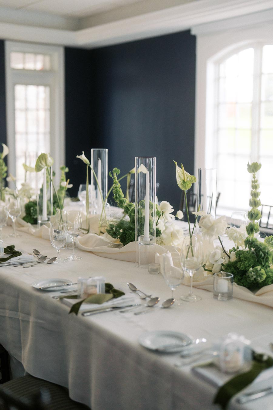 Elegant wedding reception table setting with white tablecloth, green and white floral centerpiece, tall candles, and place settings in a sophisticated venue.