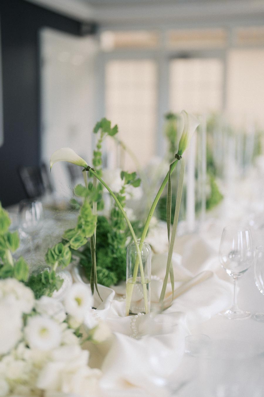 Elegant wedding table setting featuring white calla lilies and green foliage in glass vases, with soft focus on delicate white drapery and arranged glassware, creating a sophisticated and serene ambiance.