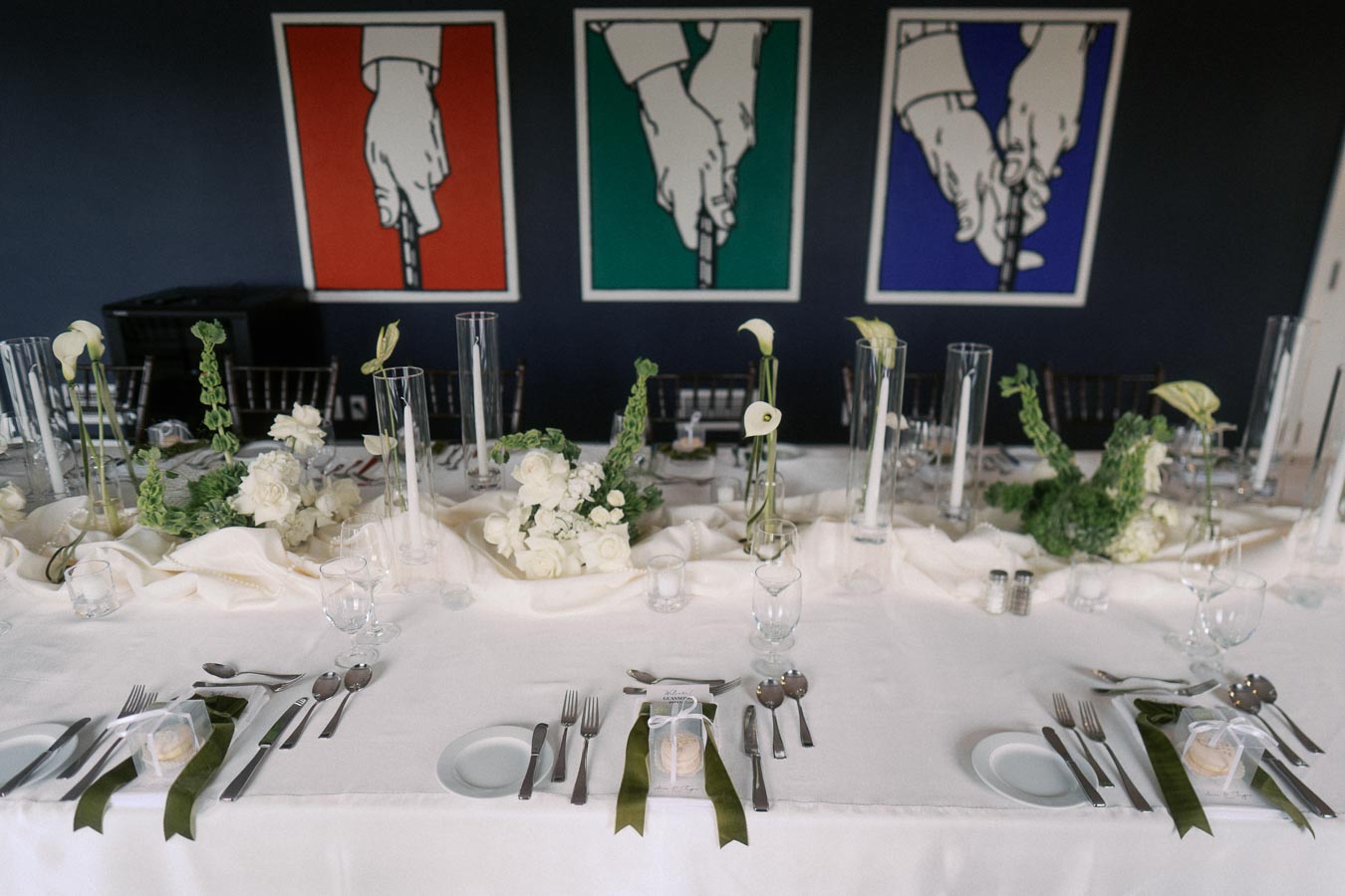 Elegantly set dinner table with white tablecloth, floral centerpieces, and modern artwork in the background, featuring minimalist calla lilies in tall glass vases and neatly arranged cutlery and glassware.