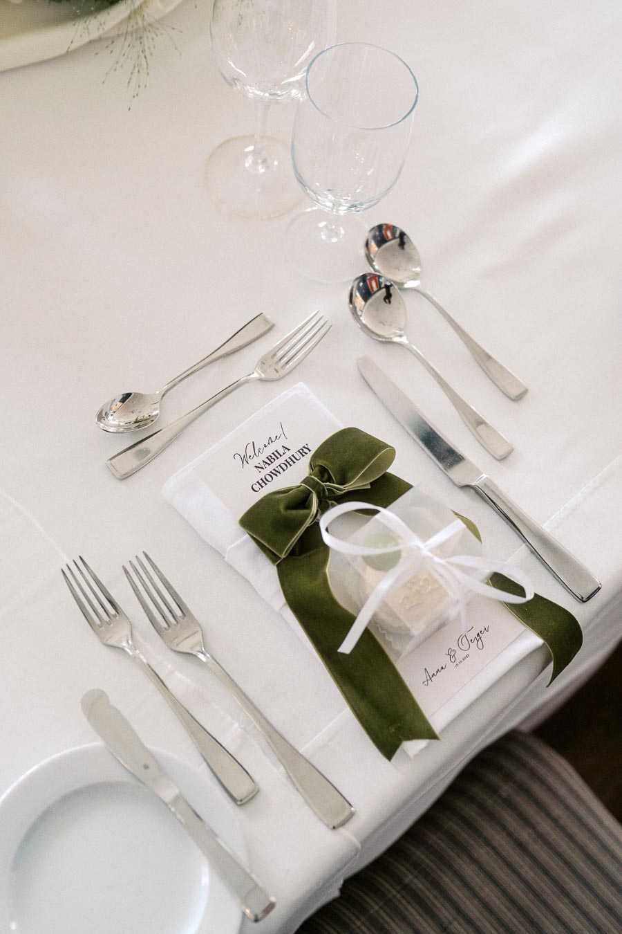 Elegant wedding table setting with cutlery and a personalized napkin featuring a green ribbon, glassware, and a small gift box on a white tablecloth.