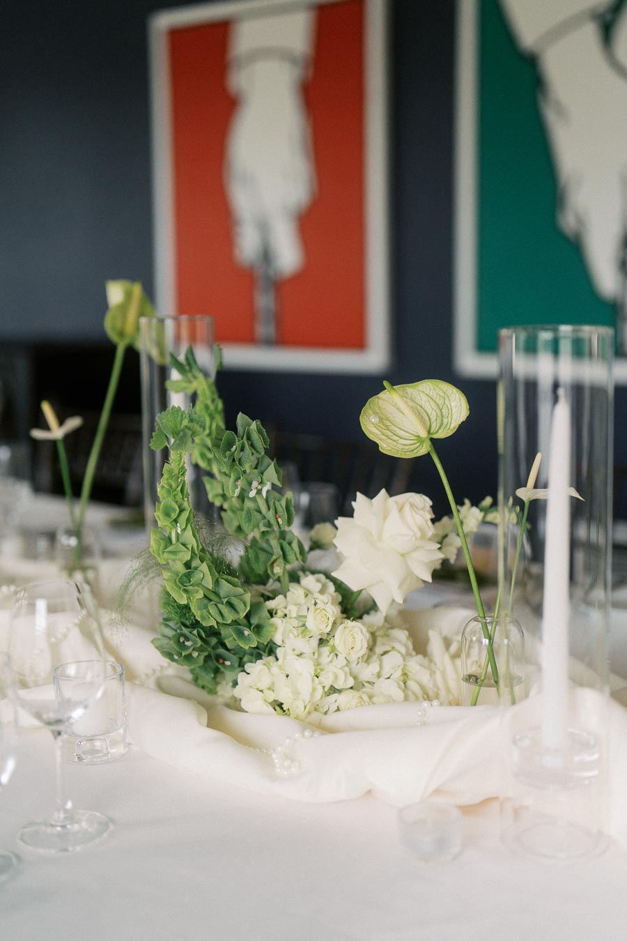 Elegant dining table setup with white floral centerpiece, featuring roses and greenery, complemented by tall candle holders and glassware; modern art in background. Perfect for showcasing sophisticated event décor.
