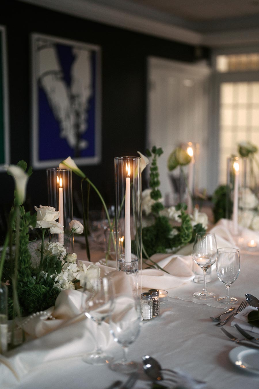 Elegant table setting for a formal dinner party featuring white floral arrangements, tall lit candles, and neatly arranged glassware on a white tablecloth.