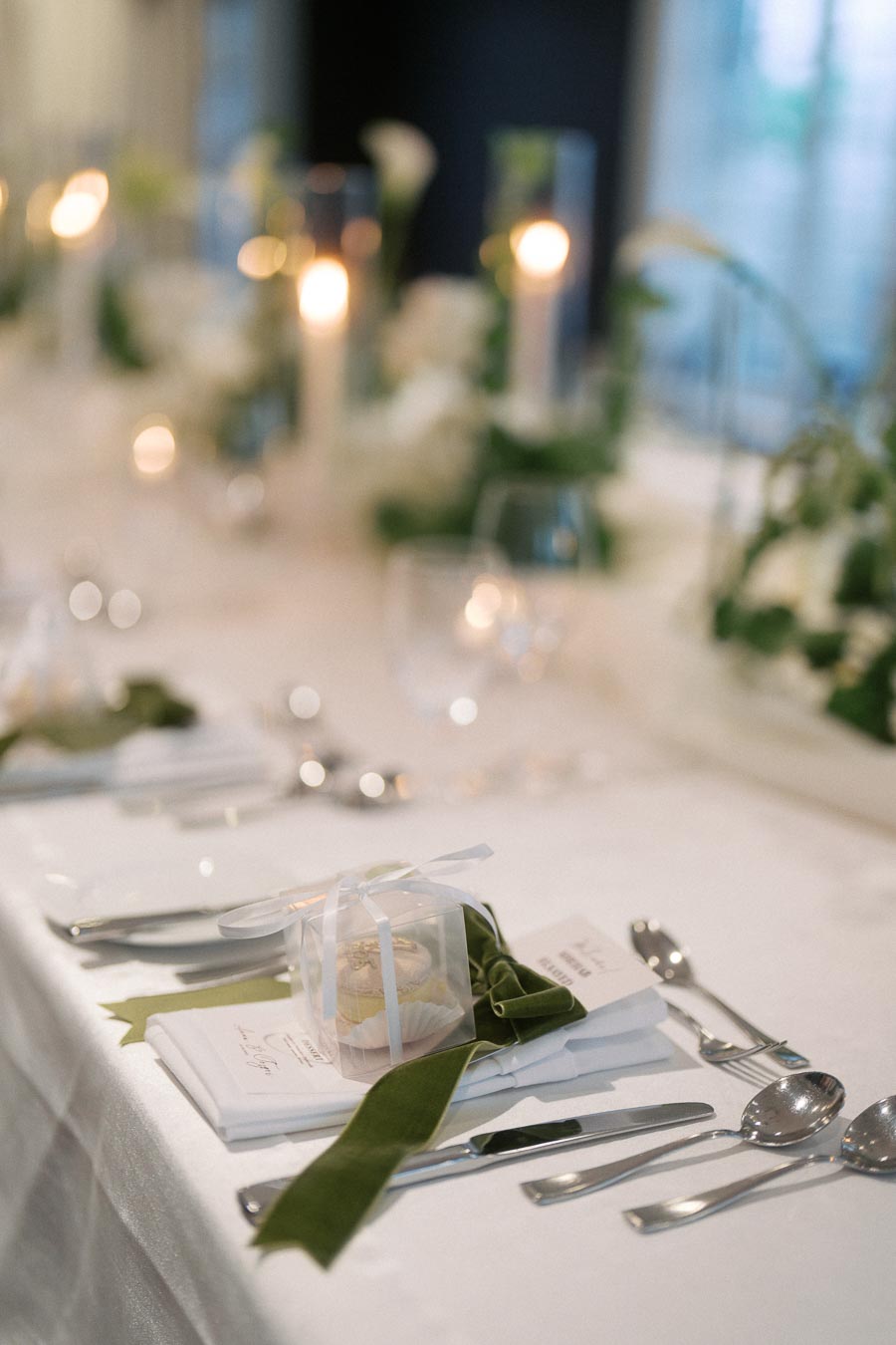 Elegant wedding table setting with white tablecloth, neatly arranged silverware, and a decorative cupcake favor in a clear box tied with a ribbon, surrounded by soft candlelight and greenery in the background.