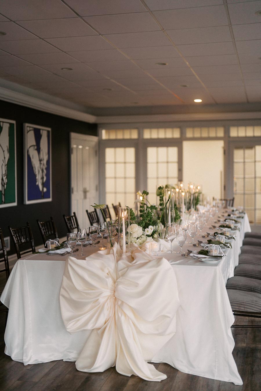 Elegant wedding reception table setup with white tablecloths, large decorative bow, and green floral centerpieces, surrounded by neatly arranged glassware and plates.
