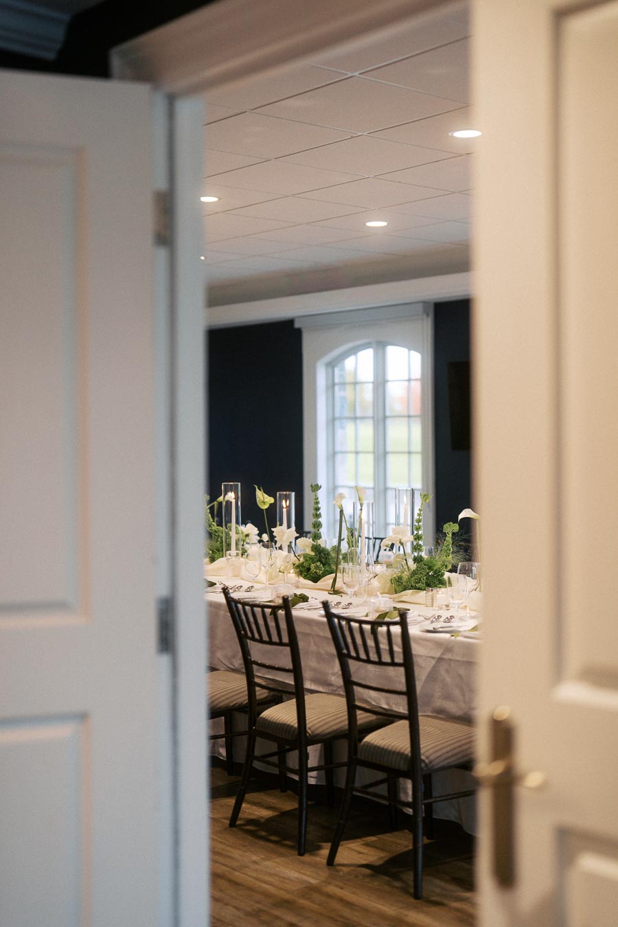 Elegant dining room with a beautifully set long table, featuring white linens and green floral centerpieces, viewed through an open door.