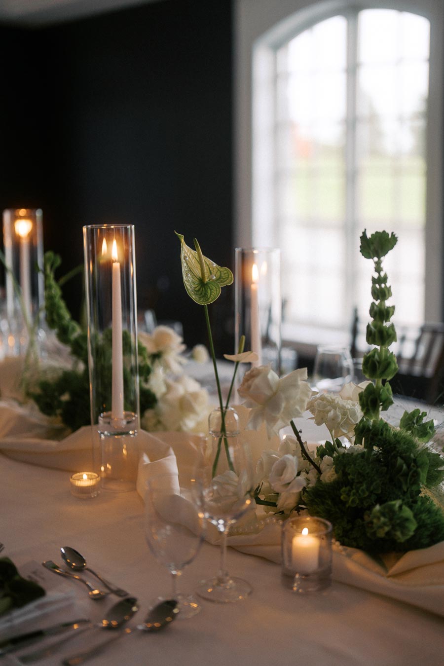 Elegant wedding table setting with tall candles, green and white floral arrangements, and soft lighting in a modern venue with large windows.