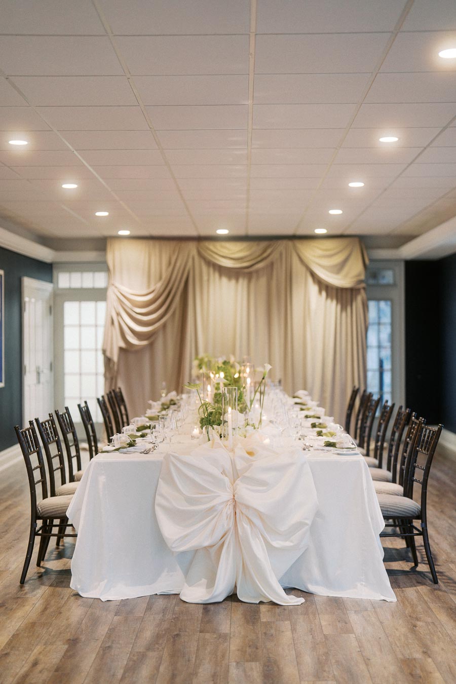 Elegant dining table setting with white tablecloth, decorative bow, and green centerpieces in a sophisticated banquet hall with soft drapery and ambient lighting.