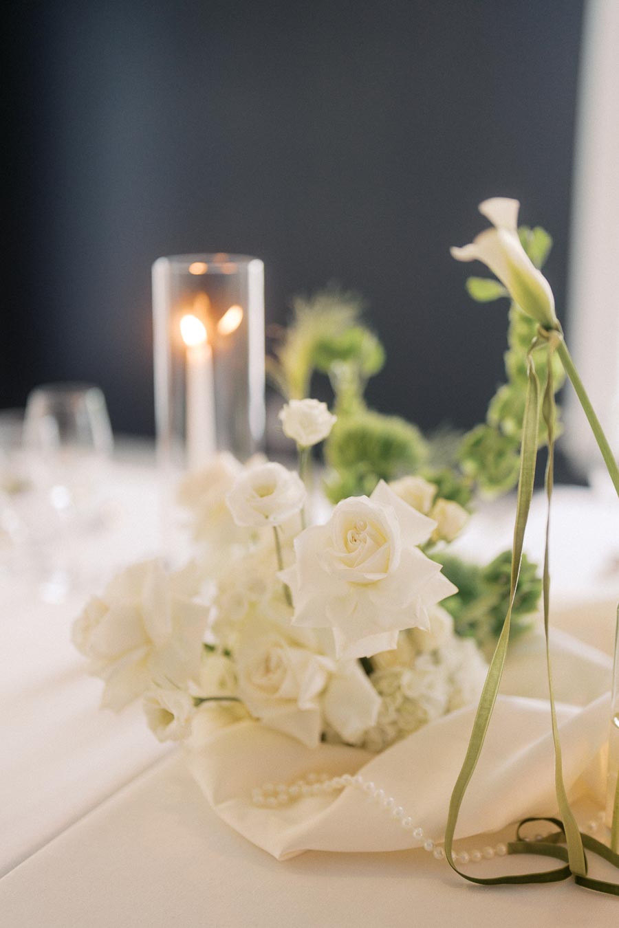 Elegant white floral centerpiece with roses, greenery, and a lit candle on a table, creating a romantic and sophisticated ambiance.