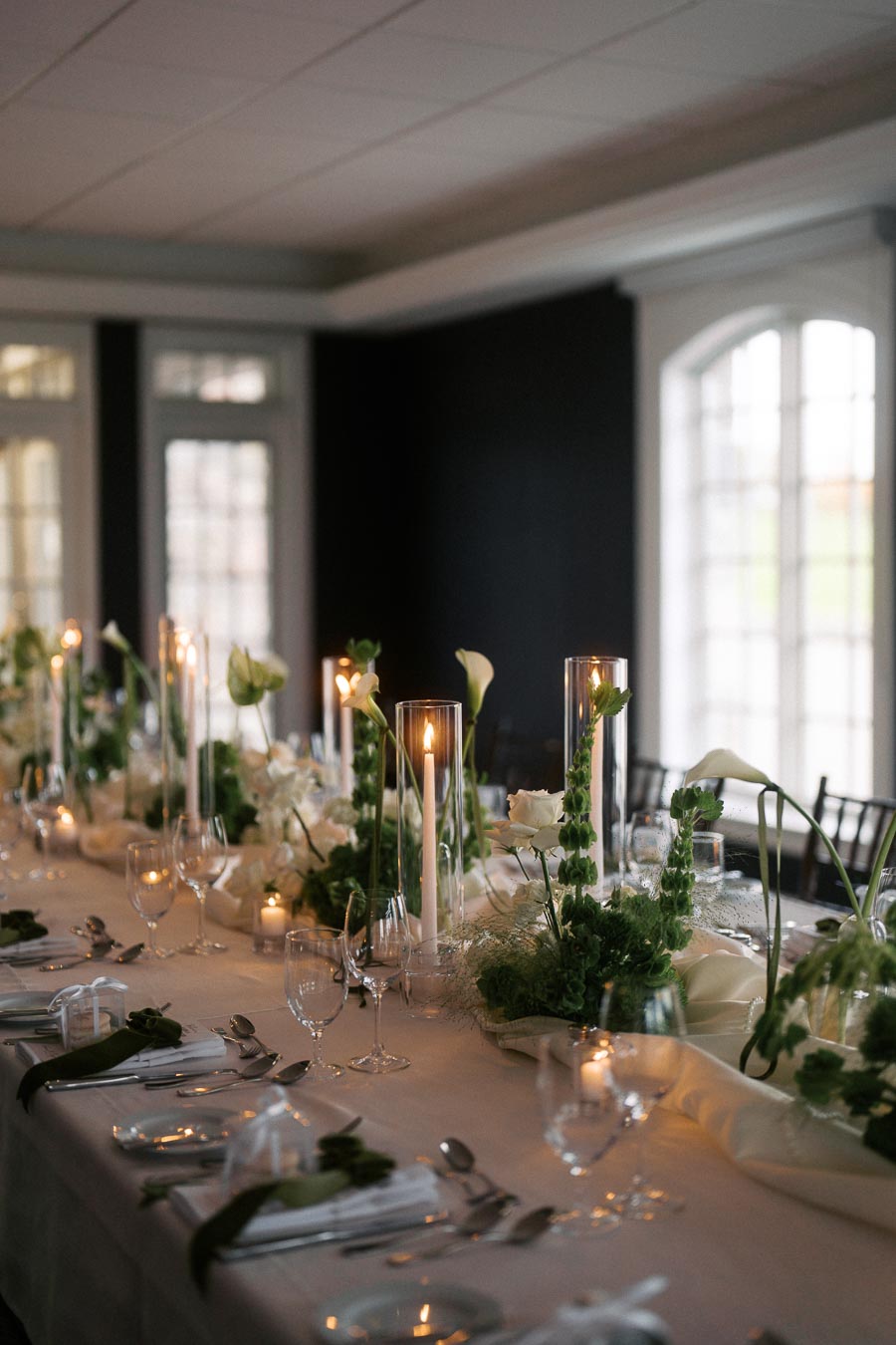 Elegant dining table setup with white floral arrangements, tall candles, and glassware in a softly lit room, ideal for weddings or special events.