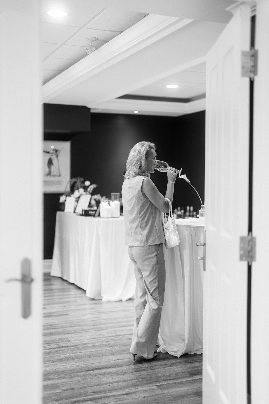 Black and white image of a woman tasting wine in an elegantly decorated room with a table set for an event, emphasizing a classy atmosphere.