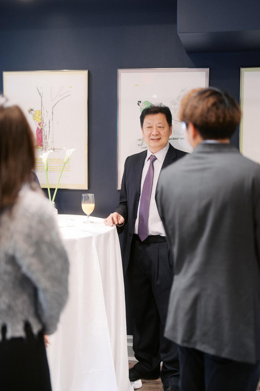 A group of people engaged in conversation at an art gallery event, with a man in a suit standing near a high table with framed artworks in the background.