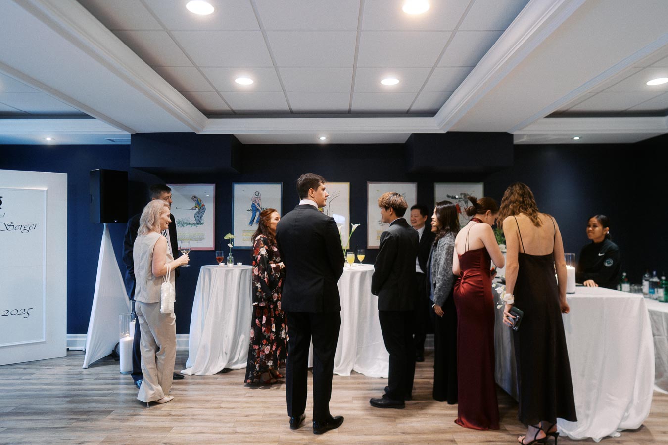 Group of elegantly dressed people socializing at a formal indoor event with a bar and artwork in the background.