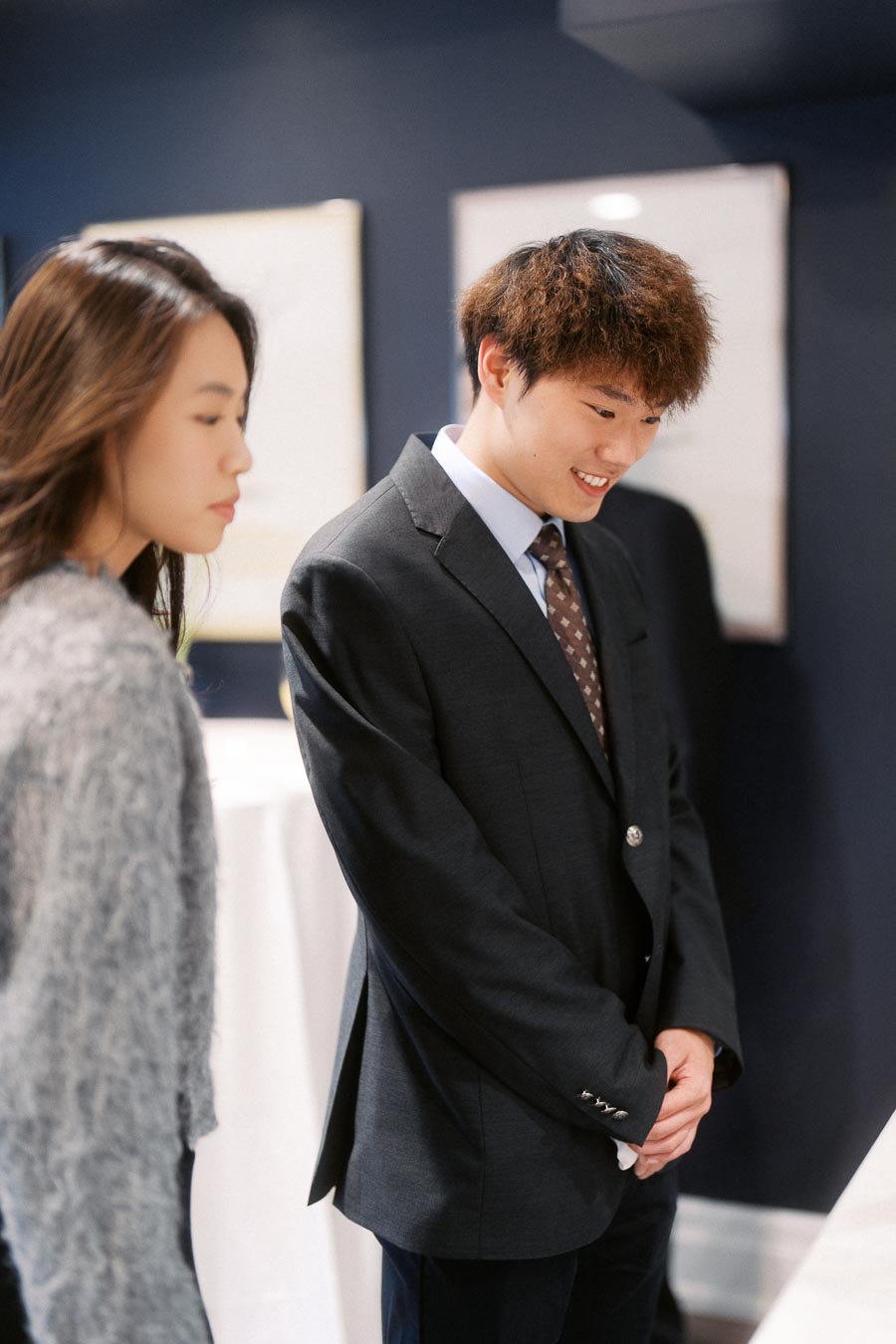 A young man in a formal suit stands smiling next to a woman wearing a textured sweater in a modern, well-lit room with blue walls and framed art.
