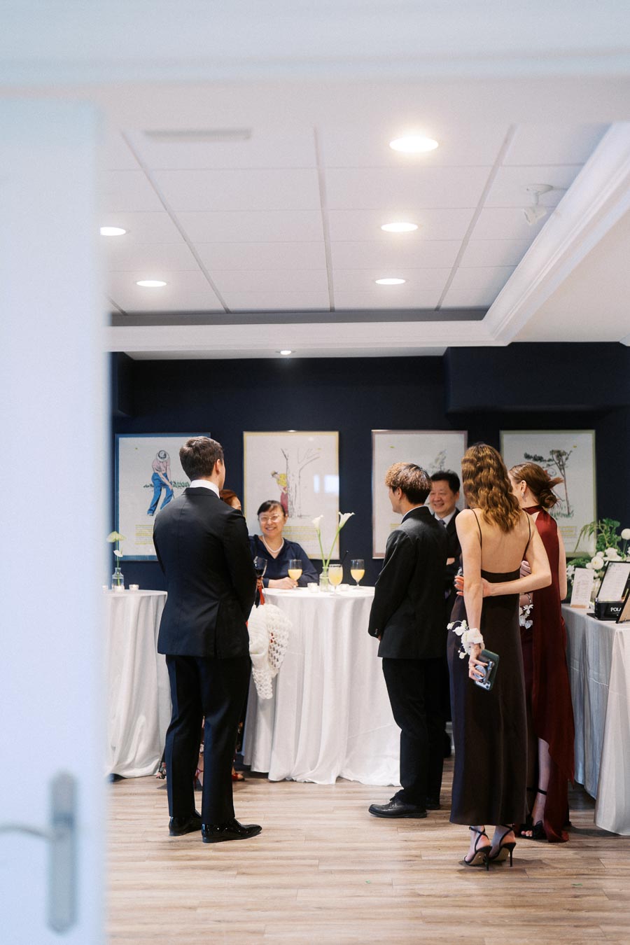 Group of elegantly dressed people socializing at a formal event, standing around a cocktail table with white tablecloths and floral decorations, in a stylishly decorated room.