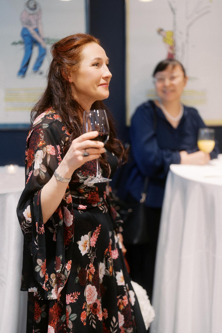 A woman in a floral dress holding a glass of red wine, smiling at a social event with another person in the background, under soft indoor lighting.