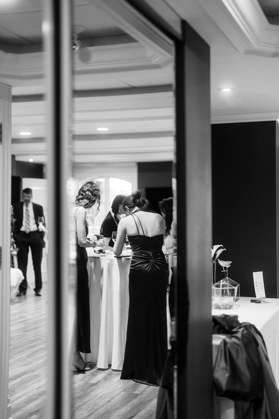 Black and white photo of elegantly dressed people at a formal event, standing around a table with drinks and decor.