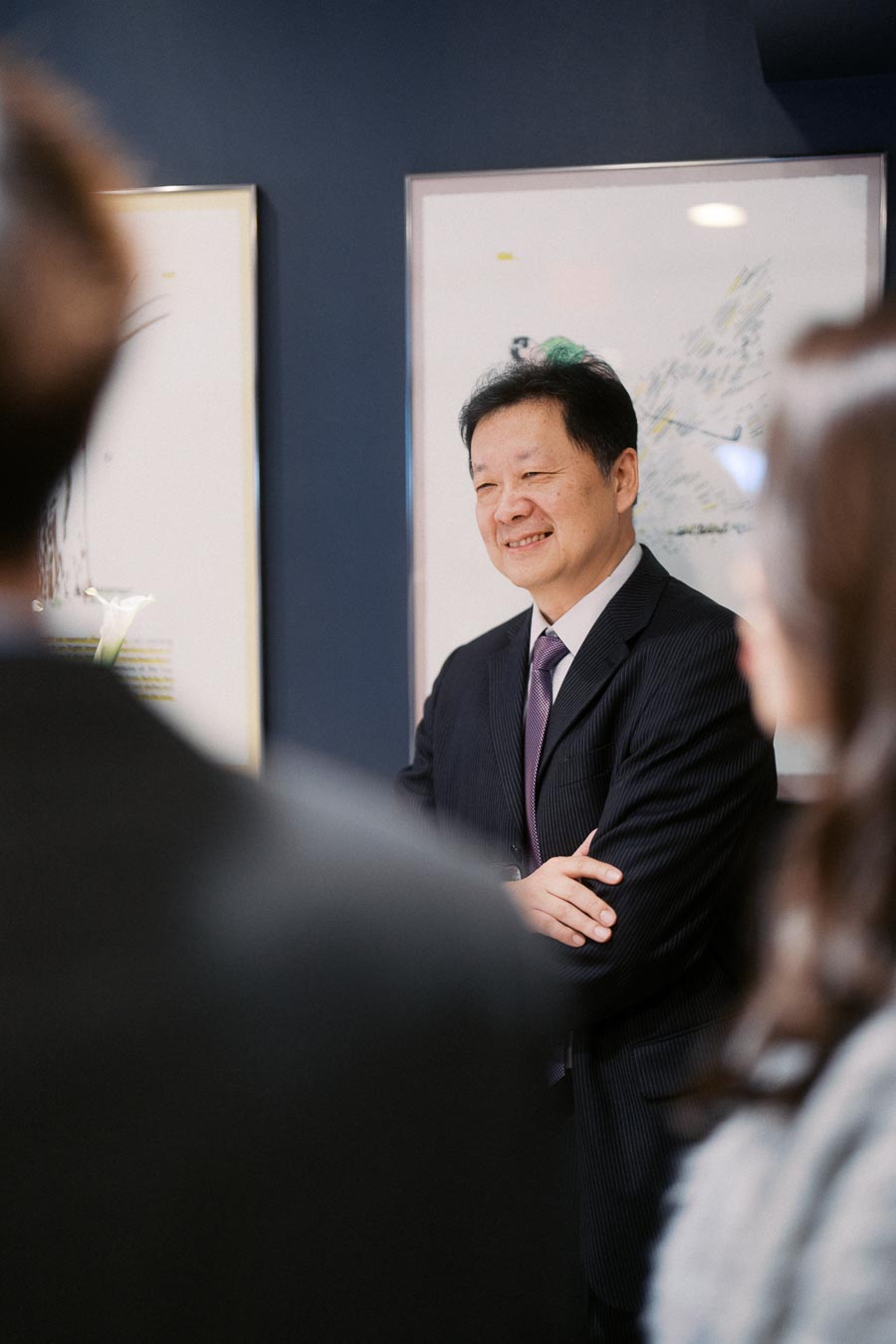 A smiling man in a suit stands confidently during a business meeting, with blurred colleagues in the foreground, set against a backdrop of framed artwork on a dark wall.
