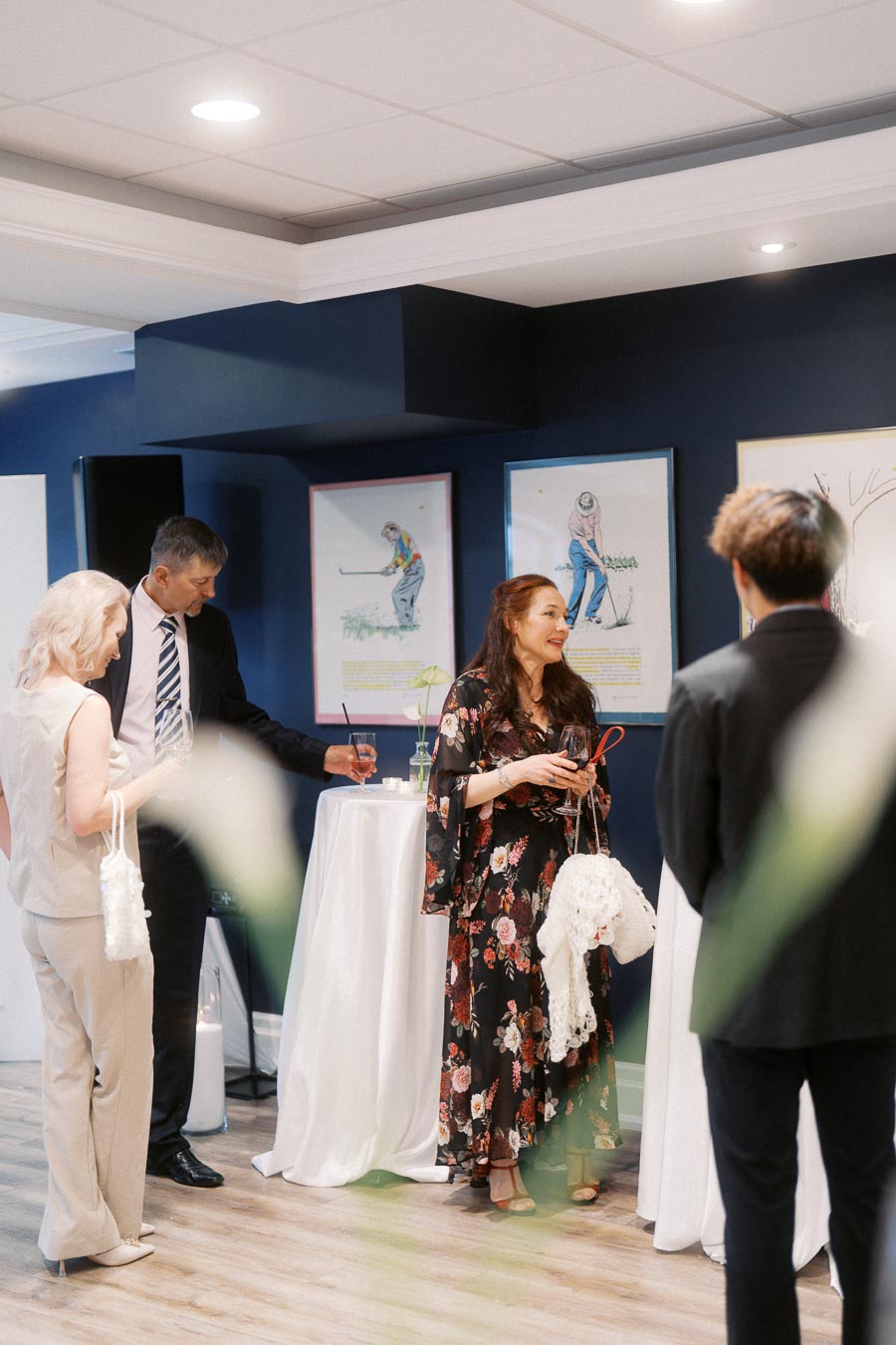 A group of elegantly dressed individuals socializing at an indoor cocktail event, standing near a small table with drinks, surrounded by golf-themed artwork on the walls.