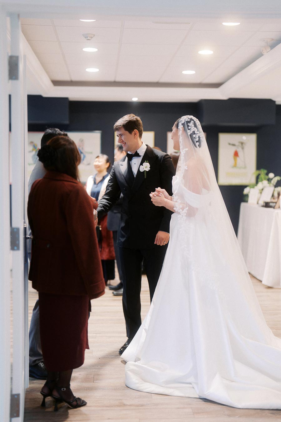 A bride in a white gown and veil stands with a groom in a black tuxedo, greeting guests in a warmly lit room decorated with framed artwork, capturing a joyful wedding reception moment.