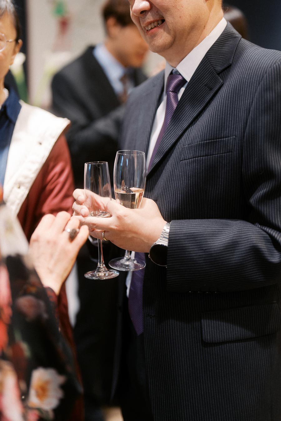 Man in a suit holding champagne glasses at a formal event, engaging in conversation with other guests.