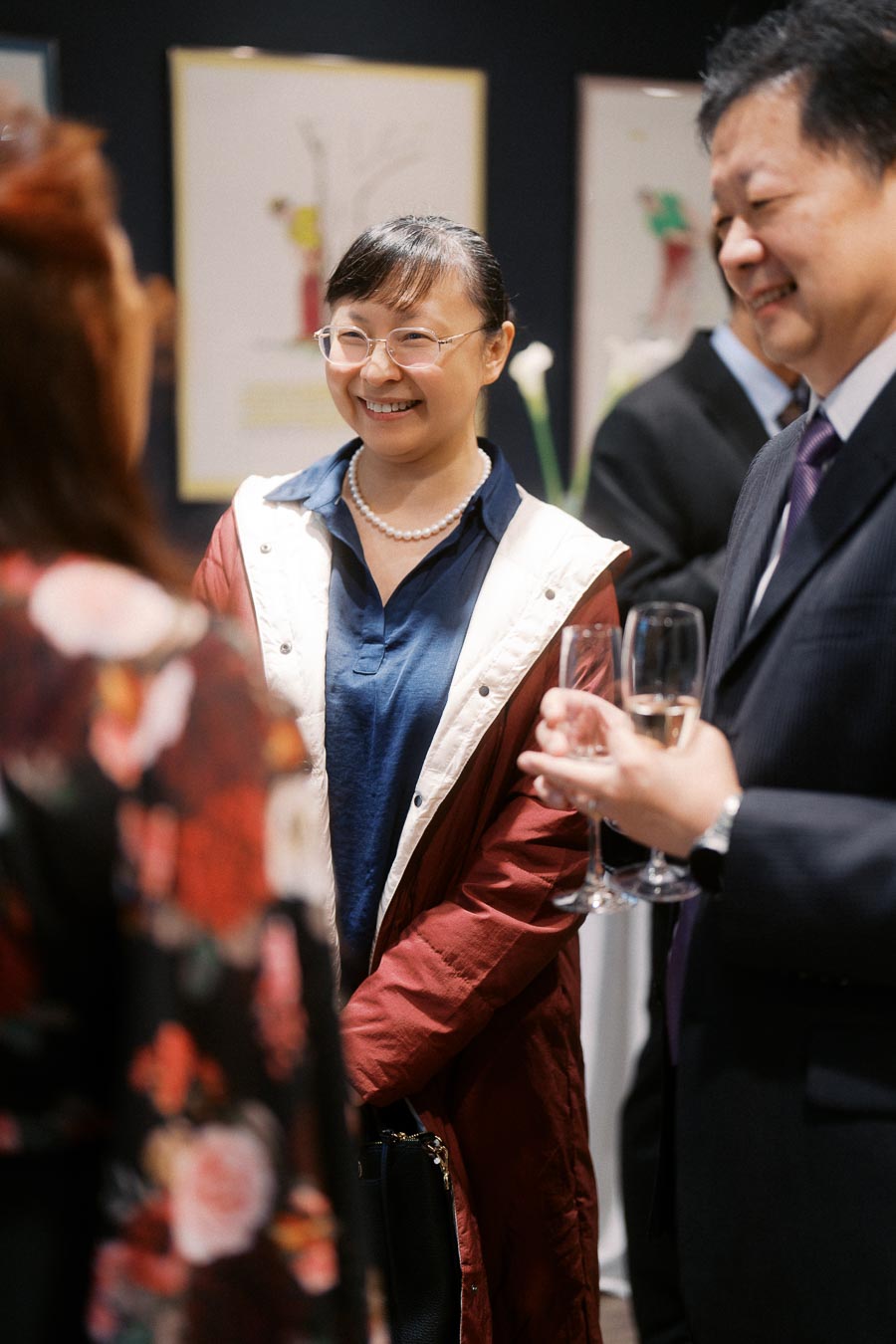 A group of people conversing at an art gallery event, one woman smiling and holding a drink, surrounded by colorful artwork and elegant attire.