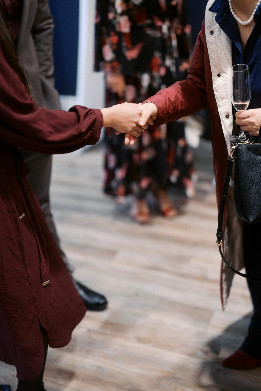 Two individuals shaking hands at a networking event, one holding a champagne glass and wearing a black floral dress, the other in a dark coat, symbolizing professional connection and collaboration.