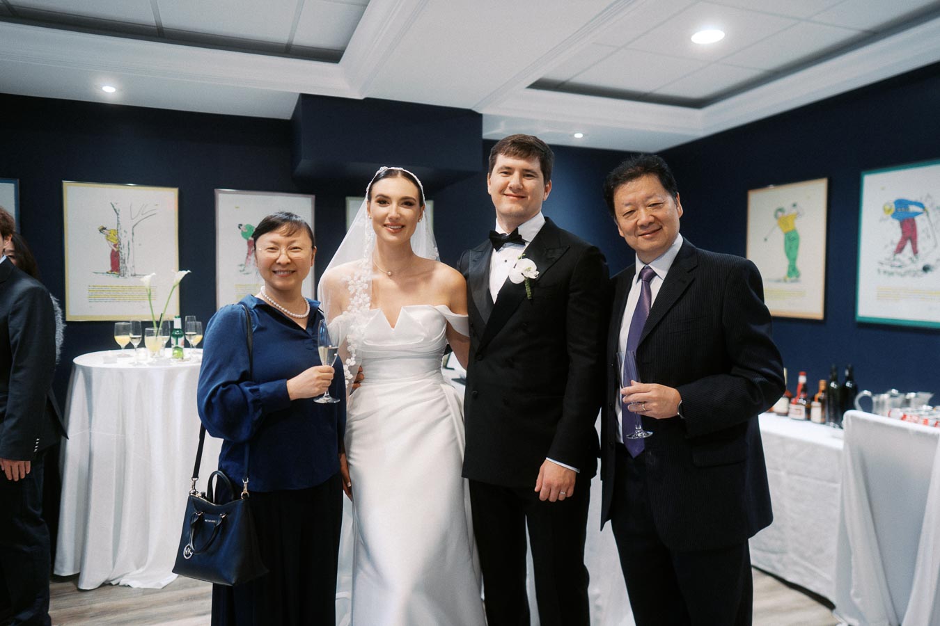 A bride in a white wedding dress and veil stands beside a groom in a black tuxedo. They are joined by two guests, one woman in a blue dress and a man in a suit, each holding champagne glasses, at a wedding reception with tables and artwork in the background.