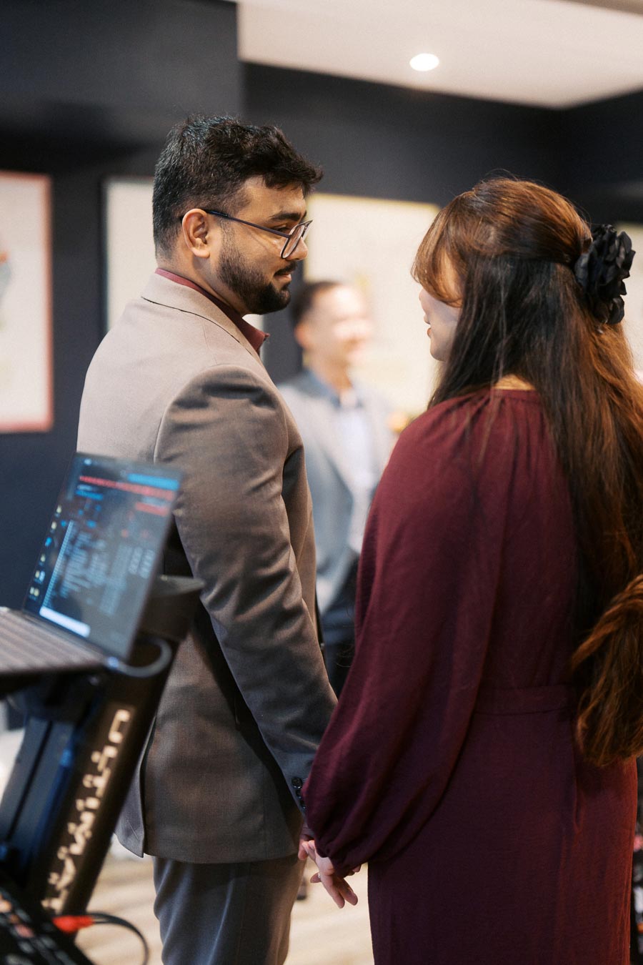 Man and woman in formal attire holding hands at an indoor event, with a laptop in the foreground and another person blurred in the background.