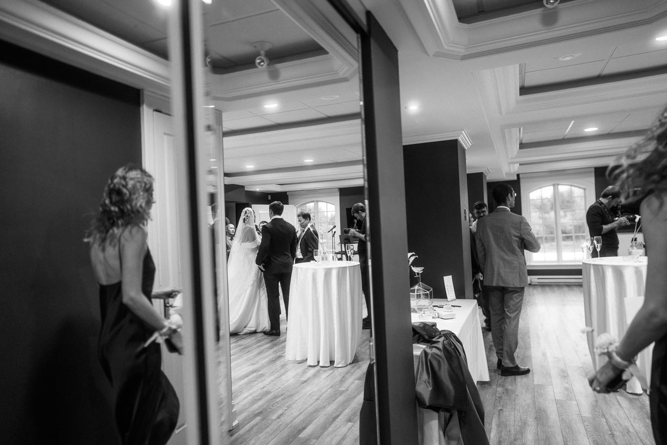 Black and white image of a formal indoor gathering, featuring a bride in a wedding dress surrounded by guests. Some attendees are dressed in suits and elegant attire, while others engage in conversation and photography around decorated tables.