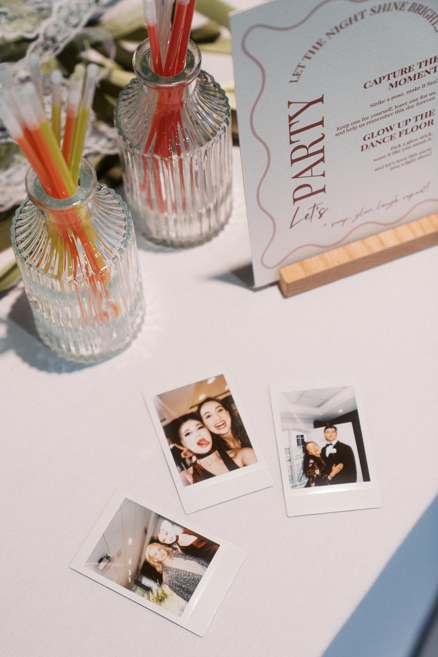 Table setup for a party featuring a decorative sign, glass jars with colorful glow sticks, and three instant photos capturing joyful moments.