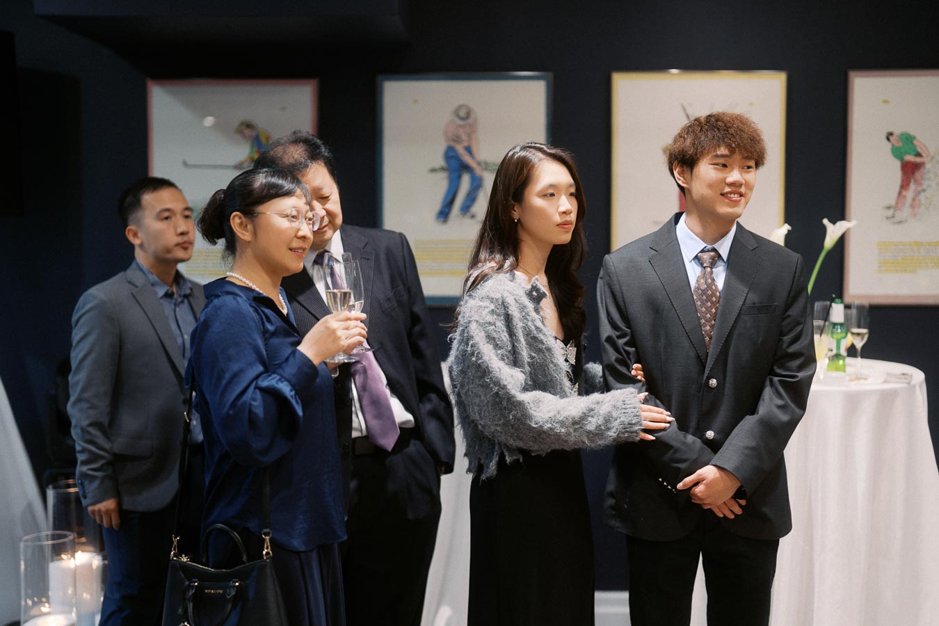 A group of well-dressed people attending an elegant indoor gathering, standing near a cocktail table with drinks, art on the walls in the background.