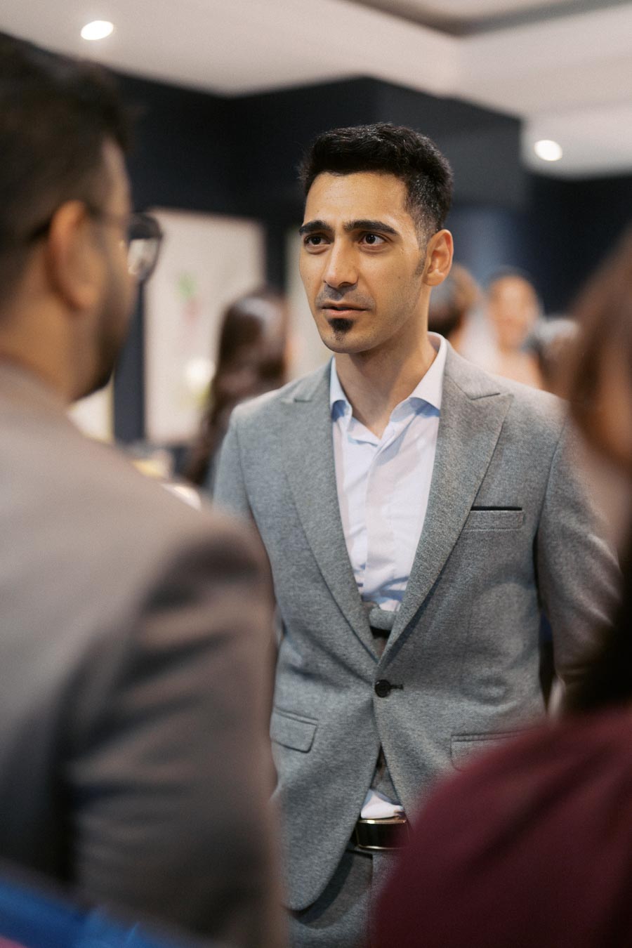 A professionally dressed man in a grey suit attentively engaging in a conversation at a social or business event, with a blurred background of people in conversation, highlighting networking and professional communication.