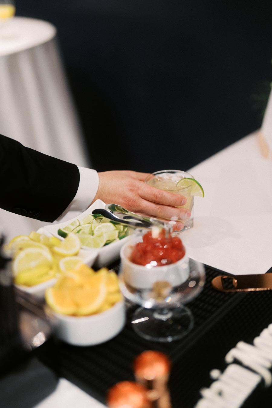 A hand in a suit reaching for a cocktail garnished with lime, surrounded by bowls of fresh red cherries and sliced lemons and limes on a bartending counter at an upscale event.