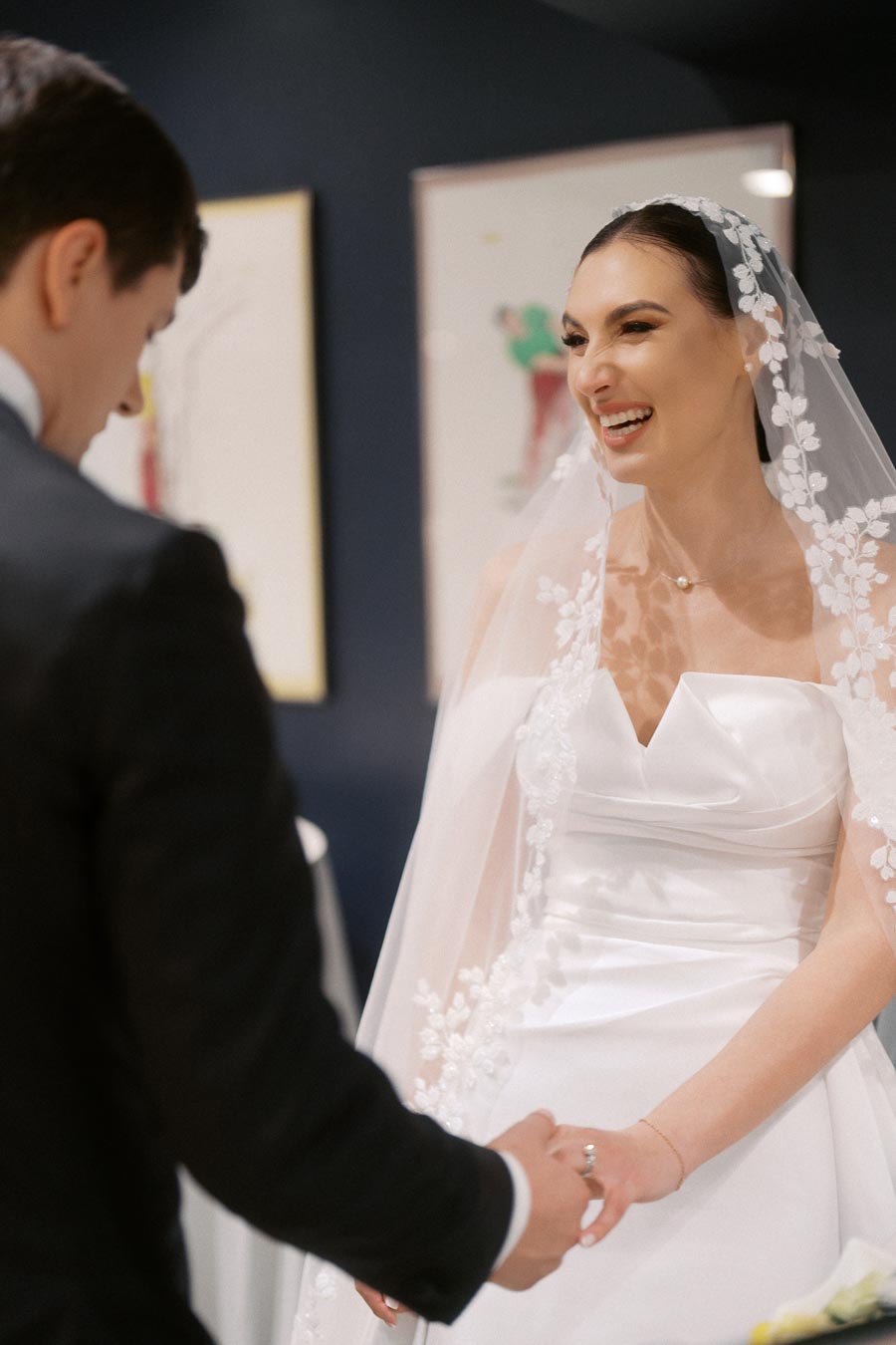 Bride smiling at groom during wedding ceremony, wearing a white dress with floral lace veil, holding hands, in an elegantly decorated room.