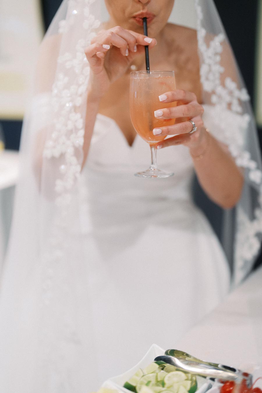Bride in white wedding dress and veil enjoying a refreshing beverage with a black straw, surrounded by a bowl of sliced limes and garnishes.