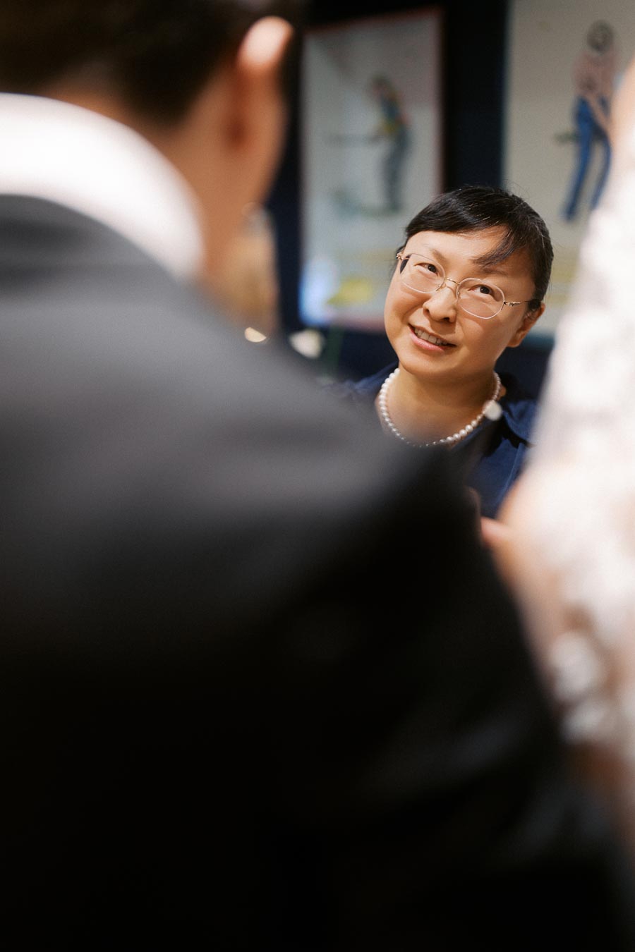 Smiling woman in a blue outfit wearing glasses and a pearl necklace, engaged in conversation at an indoor event, with blurred individuals in the foreground and colorful art in the background.